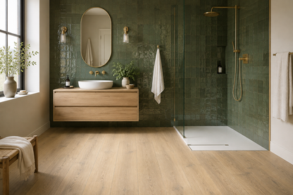 Light oak luxury click vinyl flooring in a modern bathroom with green tiled walls, walk-in shower, wood vanity and brushed brass fittings.