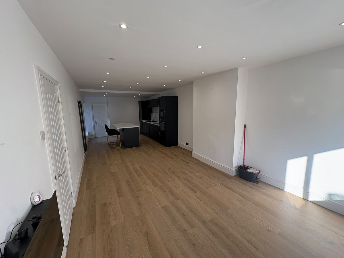 An empty modern room with light oak wood flooring, white walls, recessed ceiling lights, and an open-plan kitchen area in the background. A red-handled broom and a gray dustpan sit on the floor against the right wall.