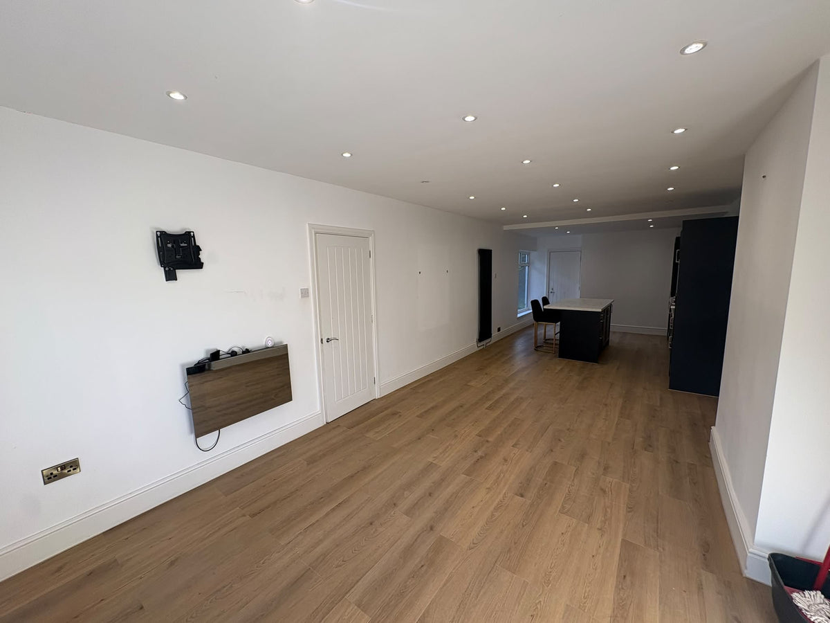 An empty modern room with light oak wood laminate flooring, white walls, recessed ceiling lights, a white door, and a black wall-mounted storage unit on the left.
