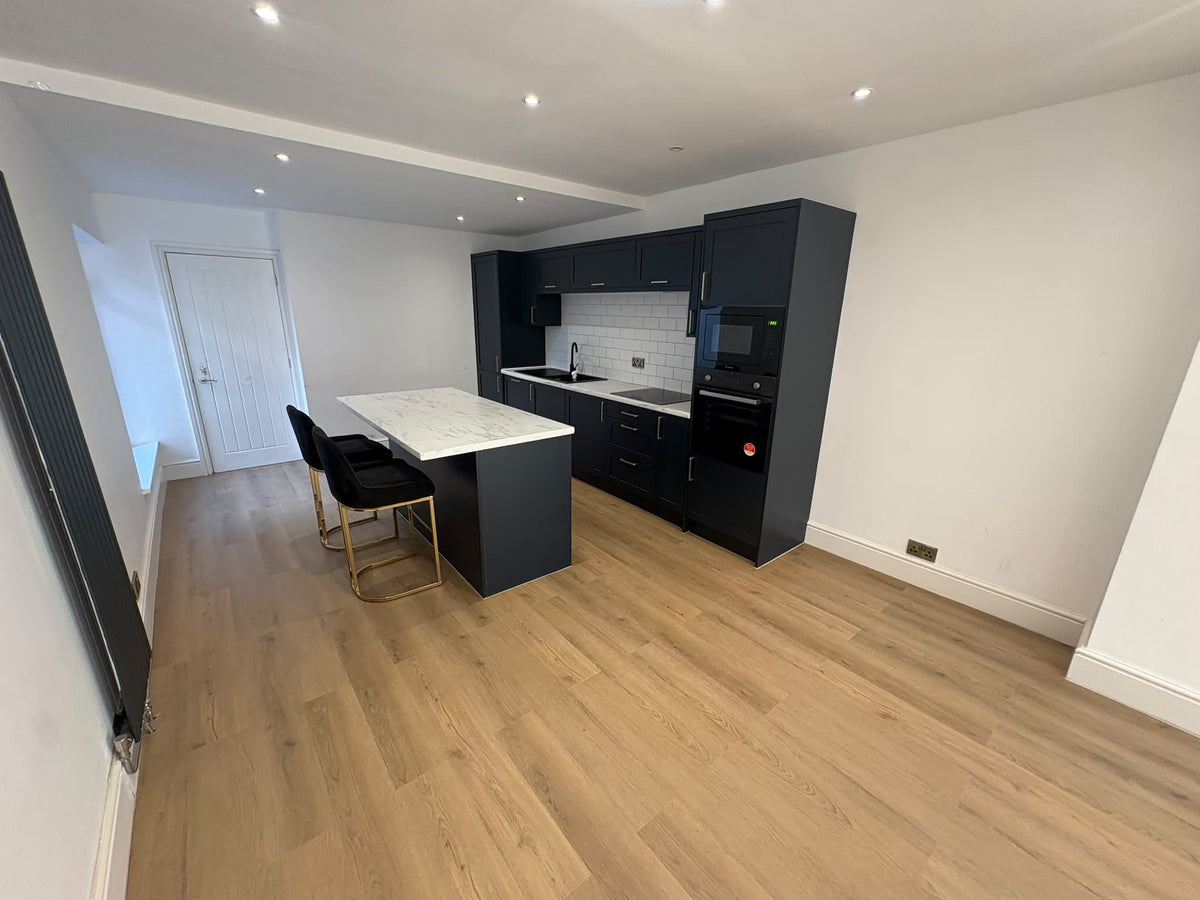 A modern kitchen island with a white marble-look top and black base cabinets, set in a bright room with light wood flooring and white walls. Two black bar stools with gold legs are placed beside the island.