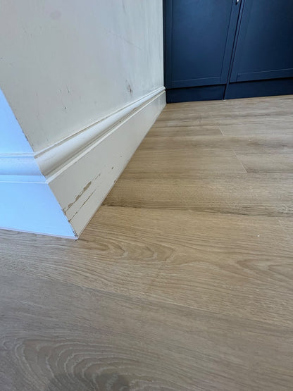 Light oak wood laminate flooring shown installed in a room corner with white baseboards and a dark blue cabinet in the background.