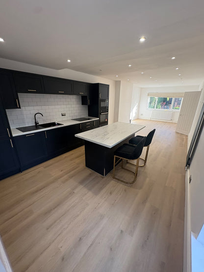 A modern kitchen interior with dark navy cabinets, a white marble-look island countertop, black bar stools with gold legs, light wood flooring, and recessed ceiling lights.
