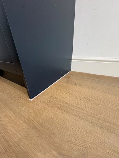 Light oak wood laminate flooring shown in a room corner with a white wall and baseboard, alongside a dark charcoal-colored cabinet.