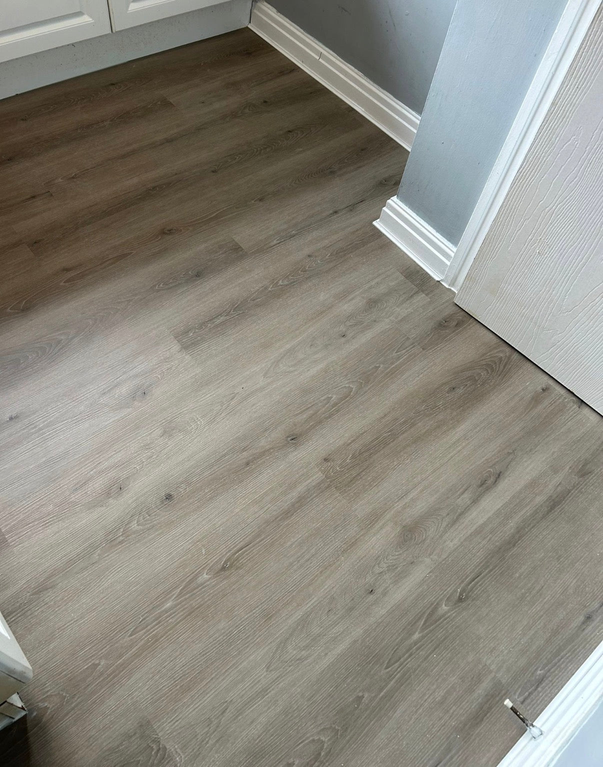 Gray-brown wood-look laminate flooring installed in a room with white baseboards and a white door frame.