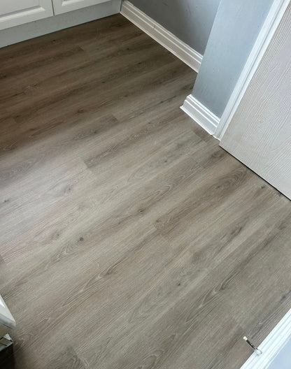 Gray-brown wood-look laminate flooring installed in a room with white baseboards and a white door frame.